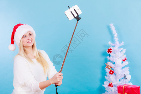 现代节日概念穿着圣诞帽子的金发女人给自己拍圣诞照用手持智能机的自拍图片
