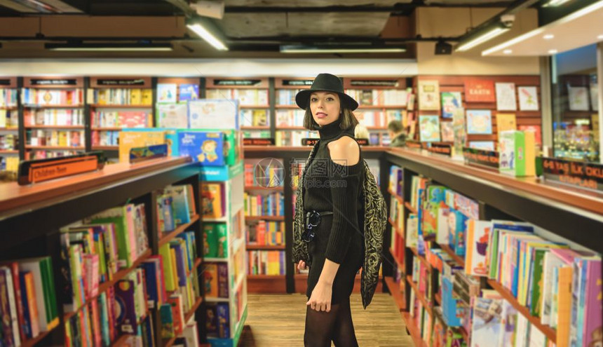 穿着黑帽子和衣服的迷人美女站在书店看着摄像机图片