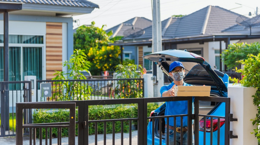 亚洲青年送货员在线亚洲青年送货员箱装蓝帽子和T恤衫他保护面罩根据宵禁检疫大流行冠状COVID19服务客户在房子门前图片