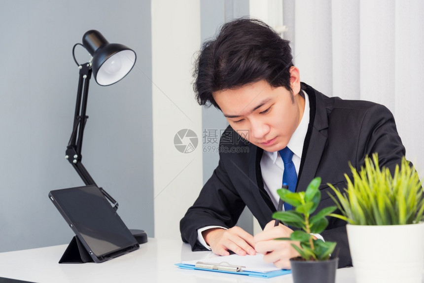 在家工作亚洲青年商人电视会议话或面对他笑着微地看用智能数字平板电脑坐在桌上的纸主办公室写笔记本资料图片