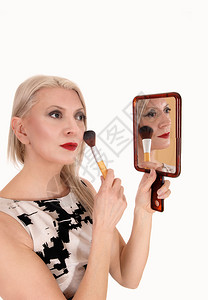 一个金发中年女子举着镜用刷修整化妆品穿着衣服与白种背景隔绝图片