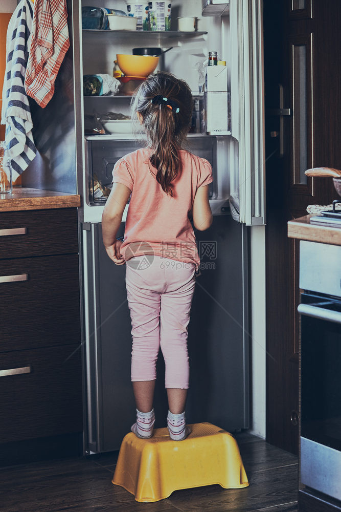 小女孩站在一个儿童凳子上在开着的冰箱前寻找食品中的冰淇淋真实人情况笑声之中正面霜图片