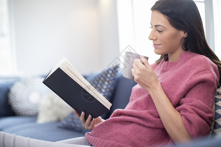 在家索法沙发上喝热饮料放松休闲的妇女阅读书保持轻松识字背景图片
