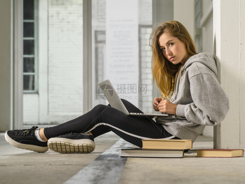 一种学习年轻的名女学生拿着笔记本电脑和书籍坐在走廊上的照片图片