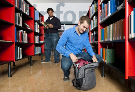 男人跪下从背包里拿东西在图书馆之间的公共图书馆里而另一个人则在背景中失去焦点笑声在室内架子大学背景图片