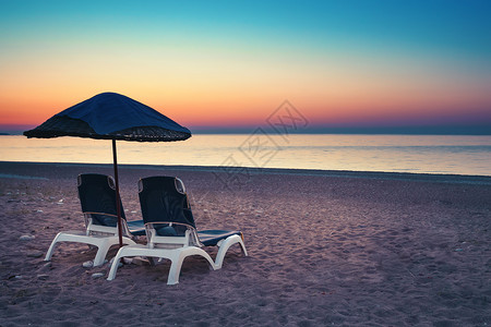 闲暇在日落的桑迪海滩沙上的伞状和两张阳床在海边和日落天空背景下带有防晒床的桑迪海滩放松和旅行的保护伞概念以及日落时在海滩上的两个背景图片