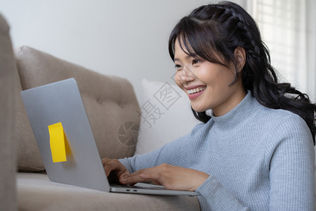 商业快乐的沙发亚裔妇女在家里使用笔记本电脑亚洲女孩在家工作时坐着笑微图片