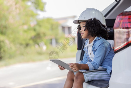 离开一个长着卷黑头发的年轻女孩指着一个工程师帽子坐在一辆梦想有天成为工程师的汽车后座上乐趣做梦背景图片