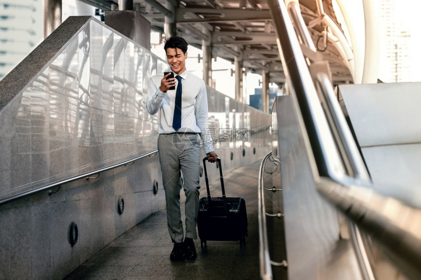 上市在机场或公共交通站使用移动电话与手提箱一起在机场或公共运输站行走时使用移动电话的微笑客乘商人现代全长的生活方式步行拉图片