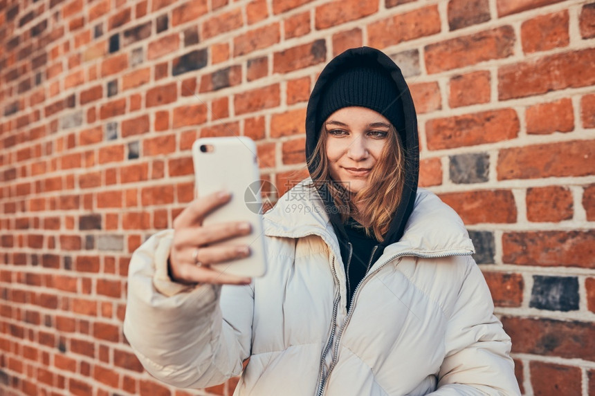 在线的年轻女子有视频电话远程交谈拍自照片手持智能机站在砖墙上松弛偏僻的图片