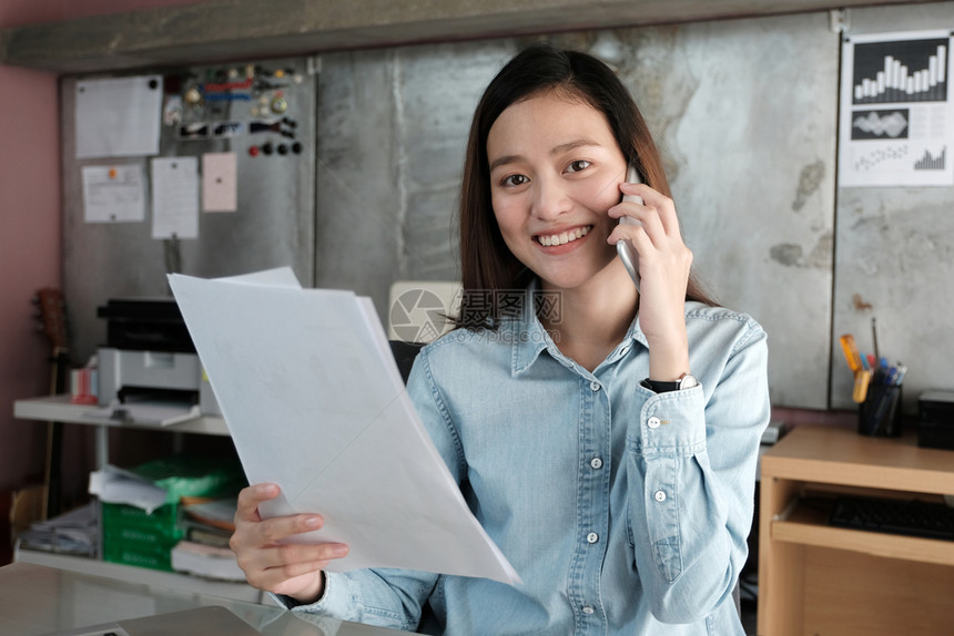 年轻的忙碌亚洲女商人在办公桌讲电话和持有文件商业临时生活方式等青年女商人手机图片