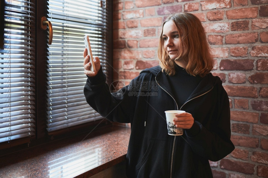 聊天在线的年轻女有视频电话远程交谈自拍照持智能手机的自拍喝咖啡放松同时在办公室休息站在窗户边上发言的女孩联系图片