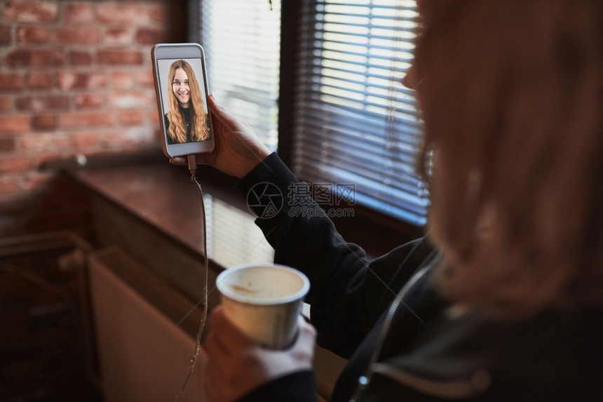 尽管保持年轻女有视频电话远程交谈自拍照持智能手机的自拍喝咖啡放松同时在办公室休息站在窗户边上发言的女孩照片图片