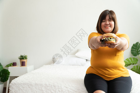 健康饥饿的超重妇女笑着拿汉堡包坐在卧室里她非常快乐享受餐吃狂食饮紊乱症概念快速地加大尺码图片
