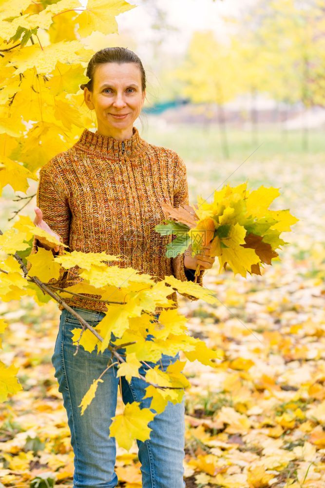 外部一个笑着的成年天主教女人秋在公园里采黄色的树叶愉快美丽图片