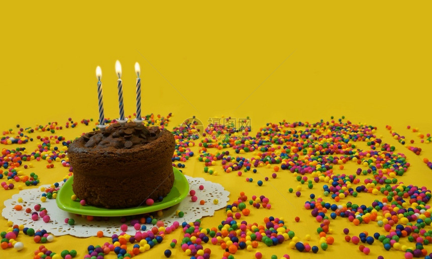红色的黄包围巧克力生日蛋糕3个蓝色和白蜡烛在绿的小盘子上点亮周围是黄色背景的糖果球有复制空间的图像图片