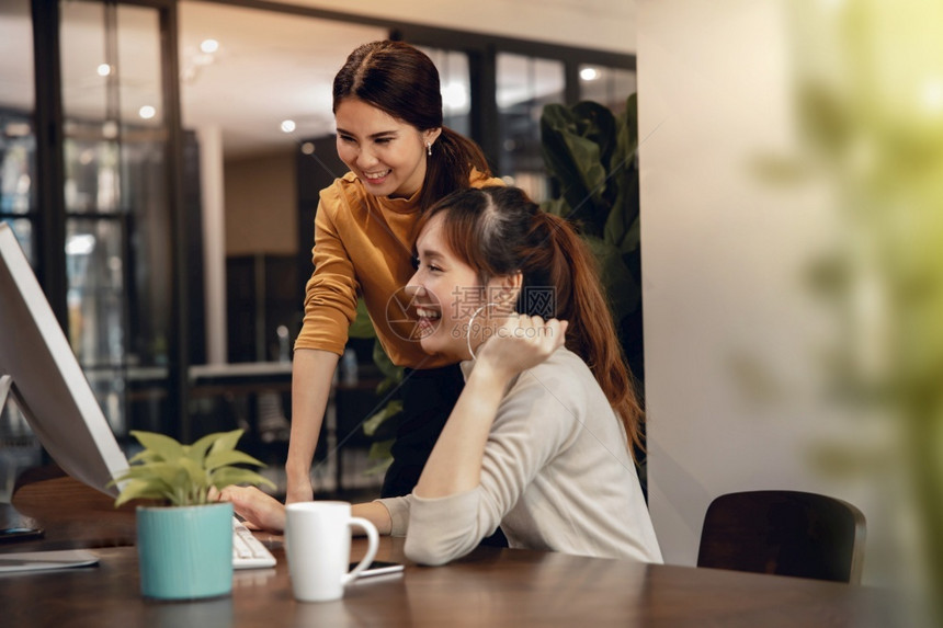 团队合作共同笑两个女商人在现代办公室计算机工作时以银幕为焦点的眼成功的员工同事图片