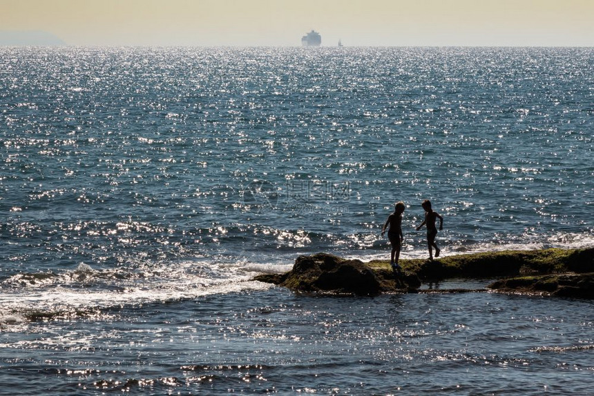 海岸线两名儿童在日落附近海礁上玩游戏的双童休月背景补给船支撑乐趣图片