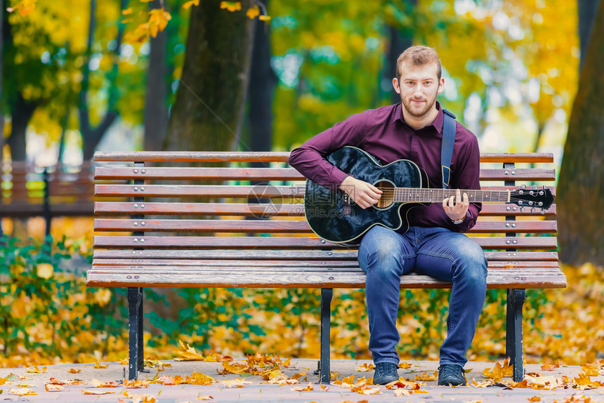 声学坐在长椅上和城市公园弹声吉他的年轻人娱乐玩图片