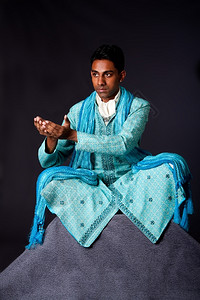 越过多蒂身穿典型民族新郎服饰的印度纯正钦丁男子坐在莲花边双腿交叉在岩石顶上伸出手来接受一份礼物上面戴着轻蓝色装饰的Dhoti和披背景图片
