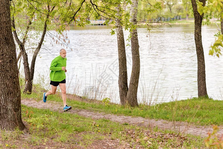 一个身着黑绿色衣服的累老年男子在一个灰色春日在靠近湖边的公园里跑来去慢者运动疲劳的背景