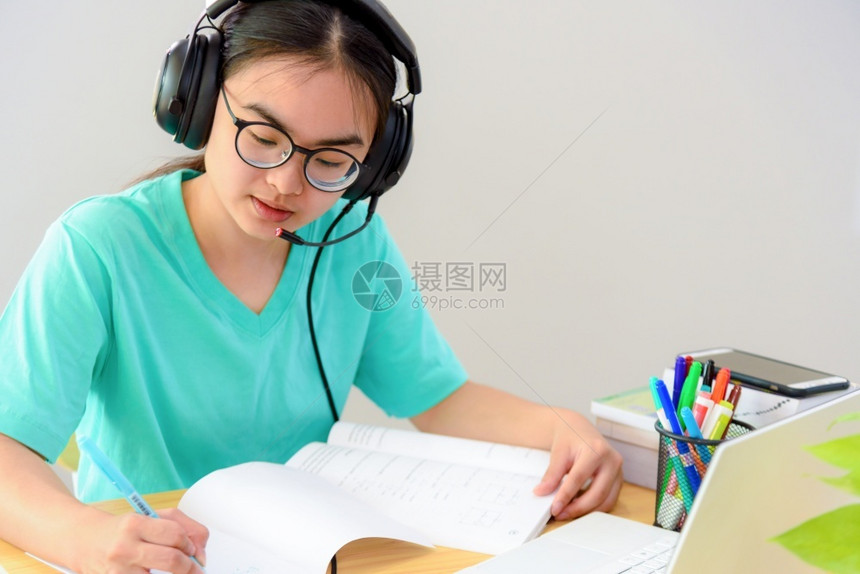 课程记录带着眼镜耳机的亚洲女学生少笑着看书本笔记电脑上的写笔记在桌子上从家中大学班级习在线研究教育亚裔妇女在书本网上学习时写笔记图片