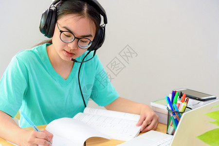 课程记录带着眼镜耳机的亚洲女学生少笑着看书本笔记电脑上的写笔记在桌子上从家中大学班级习在线研究教育亚裔妇女在书本网上学习时写笔记背景图片