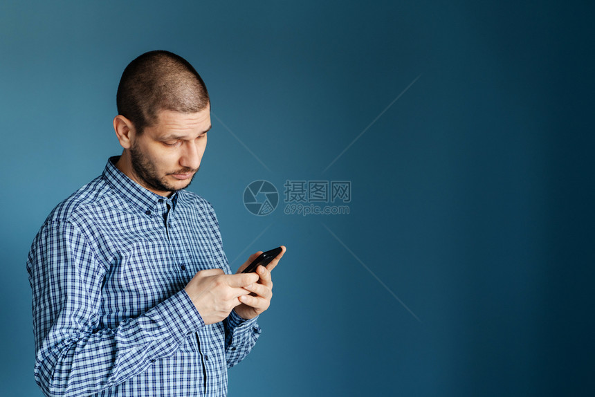 肖像白种男人穿着衬衫站在蓝背景墙前使用智能手机发送短信或浏览互联网前视图以发送信息短讯或浏览网络前视图在蓝背景墙前面穿着衬衫的白图片