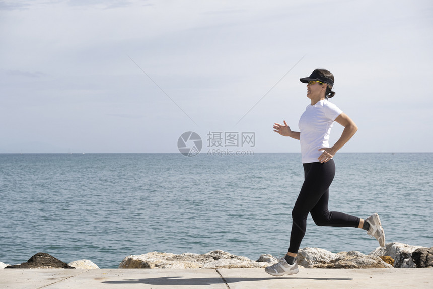 人们跑步飞跃身戴帽子和太阳眼镜的女子沿着前台奔跑背景中的大海图片