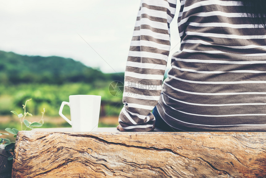 坐在年轻女人的背上坐在绿色自然山地背景的长椅上白热咖啡杯她坐着绿色自然山地背景的白色热咖啡杯背面喝香气新鲜的图片