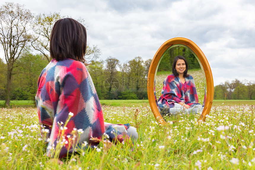 丰富多彩的公园快乐年轻科伦巴女青年在开花草地上照着镜子坐图片