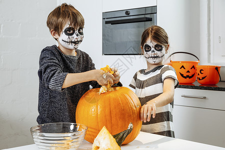 雕刻假期可怕的两个头骨脸涂油漆的小孩在厨房里刻着南瓜图片