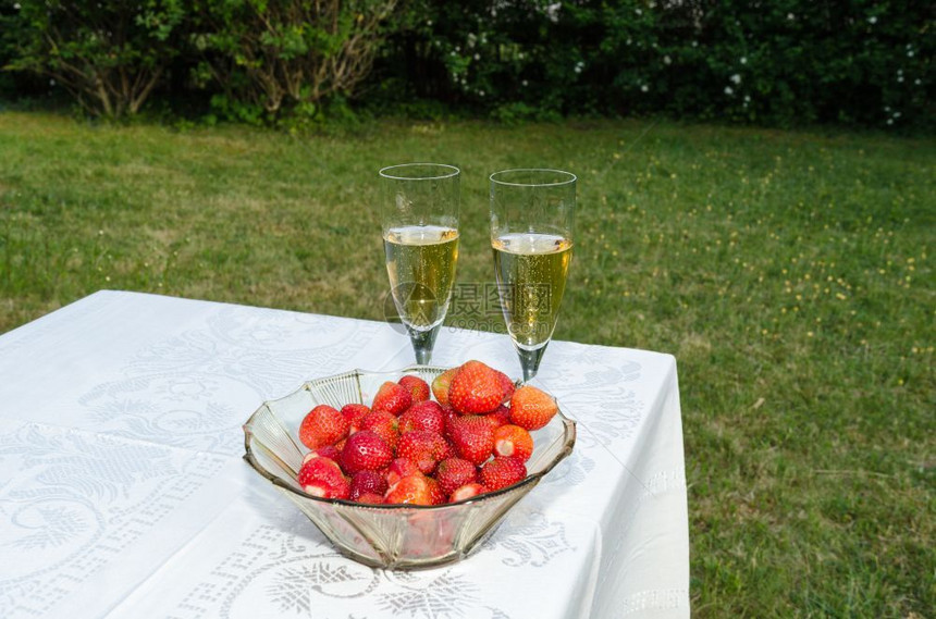 夏令周年纪念日碗加新鲜草莓和两杯玻璃在桌边上闪亮着葡萄酒花园里有白布饮料图片