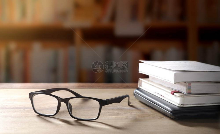 闲暇在图书馆区域背景上用照明光和模糊书架的玻璃眼镜堆叠书籍和木制桌上黑色笔记本电脑目的团体图片