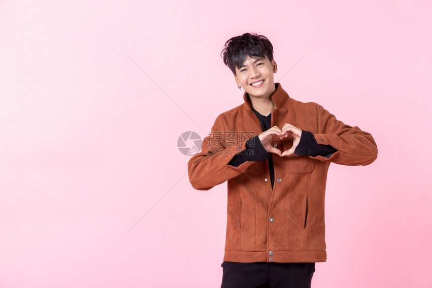 一个亚洲帅的年轻男人笑着快乐展示心形手势和侧眼看着爱中的相机在粉红空白间室背景上被孤立常设男生年轻的图片