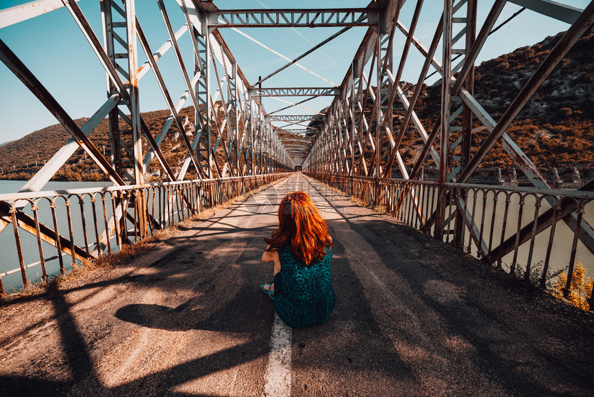 年轻的蓝色弃坐在旧钢桥上的年轻红发女青图片