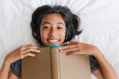 尽管亚洲小女孩笑着看镜头摄影机在床上躺着时拿棕色书籍的亚裔女孩最美景象中爱好房间背景图片