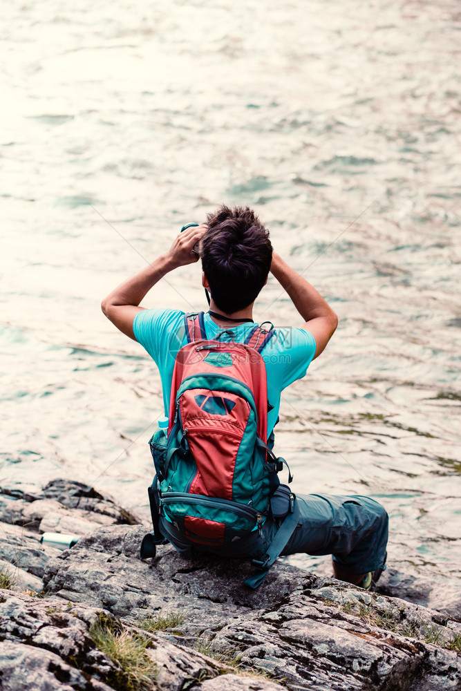男人观看白种带着背包的年轻旅游者通过望远镜看坐在河边的岩石上男孩在与背包一起游荡时度假他穿着运动服装图片