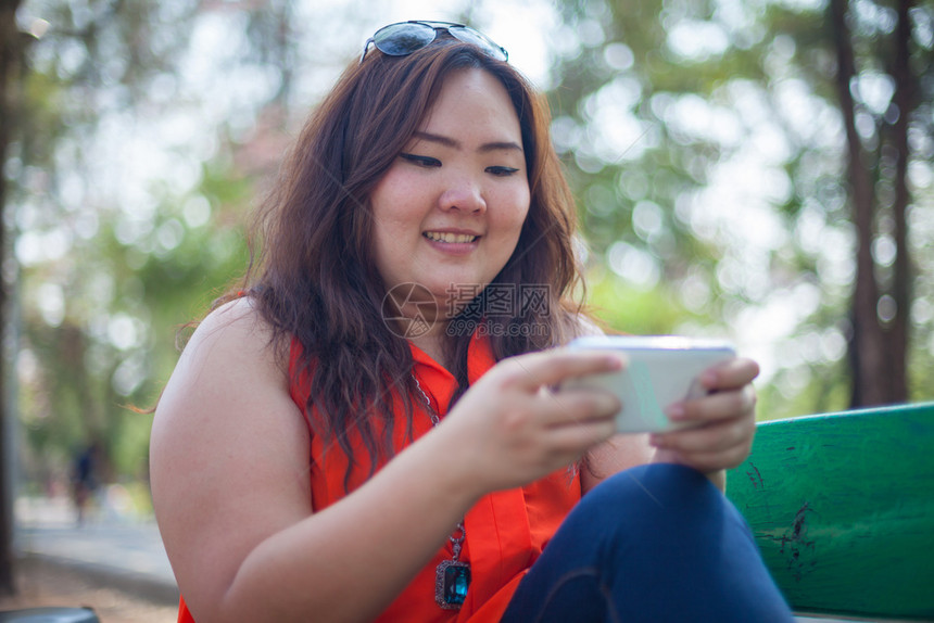 坐微笑在公园户外使用手机的快乐肥胖亚洲女人放松图片