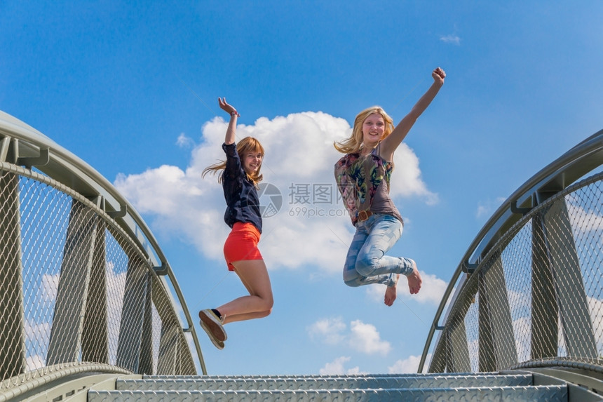 微笑正面快乐的两个和热情的少女在蓝天前跳上桥在蓝天前图片
