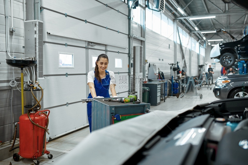 女机械师站在引擎盖上汽车服务修库穿制服的女人汽车站内部背景女机械师站在引擎盖上汽车服务室内的劳动维持图片