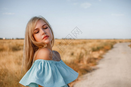 烈日炎炎头发时尚女士身着浅蓝色的轻礼服站在一个田地上旁边一条小乡村路旁有晒太阳草的年轻美丽caucasian金发美女肖像背景