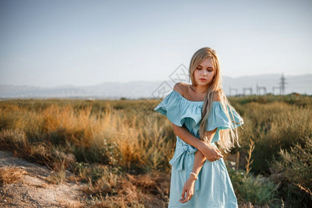 烈日炎炎常设淑女冷静的身着浅蓝色轻礼服站在一个田地上旁边一条小乡村路旁有晒太阳草的年轻美丽caucasian金发美女肖像背景
