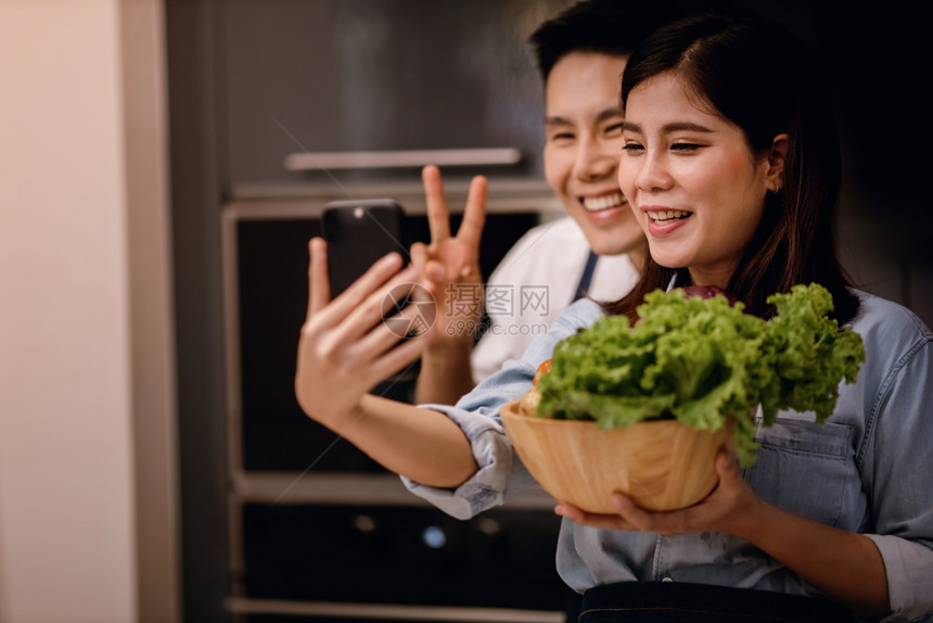 烹饪美食蔬菜一对微笑的男女在厨房里用手机自拍或通过智能手机直播同时准备素食品现代健康生活方式概念一起在家做饭图片