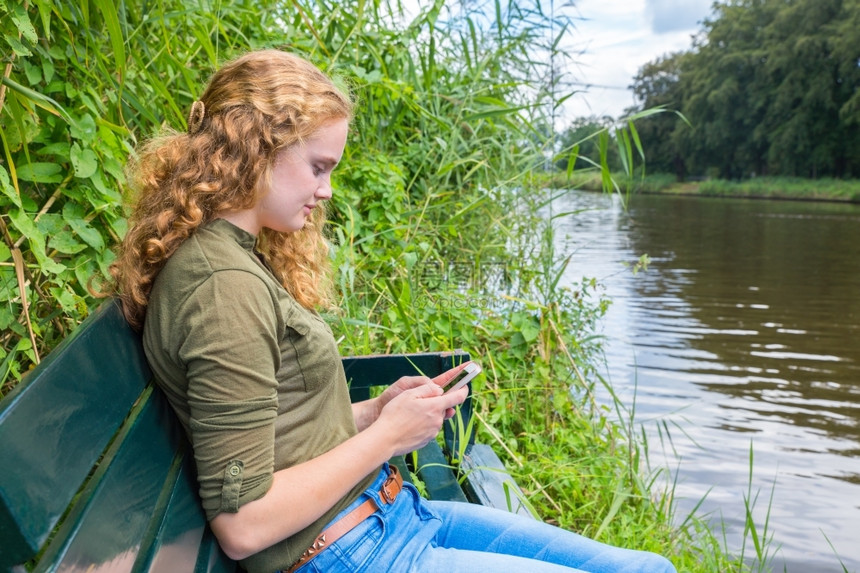 女孩年轻的在水前长椅上阅读移动电话的年轻荷兰女青手机图片