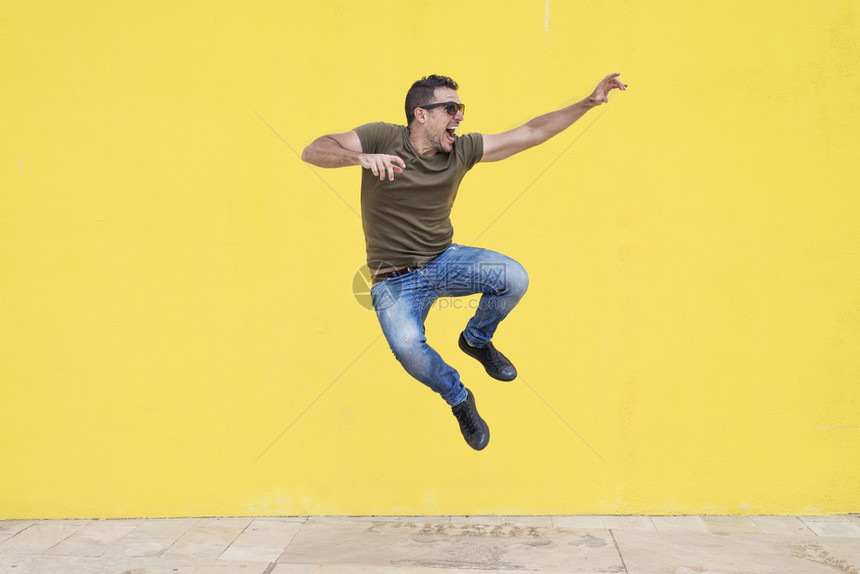全套服装带着太阳眼镜的年轻人在黄墙自由前跳跃着垂直的活力图片