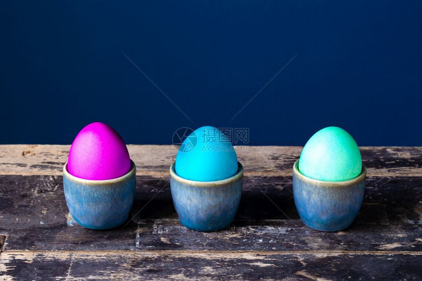 以蓝背景的鸡蛋杯在木架子上涂满彩色闪亮复活节鸡蛋手用蓝背景的鸡蛋杯在木架子上涂满彩色鲜亮复活节鸡蛋假日弹簧卡复古设计花春卡老旧设图片
