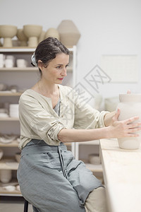 陶艺家职业器年轻女坐在桌边工作室制造粘土或陶瓷杯图片