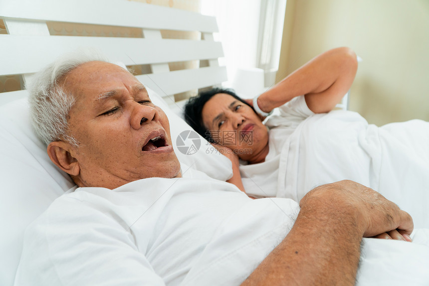 苏醒亚裔老妇人张开双眼用手遮耳盖朵躺在她丈夫旁边的床上丈夫打呼噜制造噪音婚姻问题枕头疲劳的图片