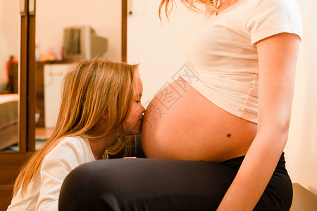 亲吻姐姐女儿在家中站立亲吻孕妇肚子等待婴儿的到来女孩在家小儿站着亲吻母肚子怀孕等待婴儿的到来未出生妈拥抱背景
